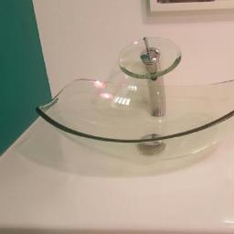 Usługi hydrauliczne - Szklana umywalka z baterią o szklanym dysku na blacie, widok z boku, na tle zielonej ściany i białej ściany z obrazem.