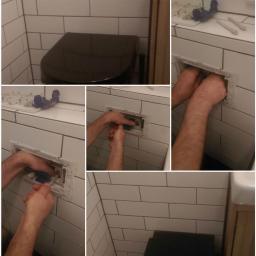 Usługi hydrauliczne - Instalacja podtynkowego systemu spłukiwania w toalecie z białymi płytkami; widoczne ręce instalatora, elementy systemu i czarna miska ustępowa w tle.