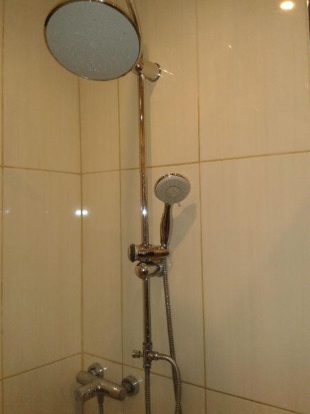 Bateria prysznicowa z deszczownicą i słuchawką, zamontowana na ścianie z jasnych płytek w kabinie prysznicowej.