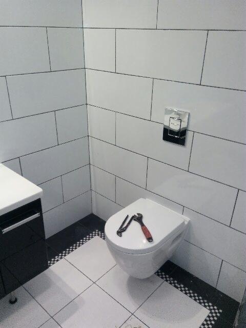 Biała, wisząca toaleta z narzędziami na desce, na tle ściany z białych płytek, widoczny fragment szafki umywalkowej i dekoracyjne wykończenie podłogi w szachownicę.