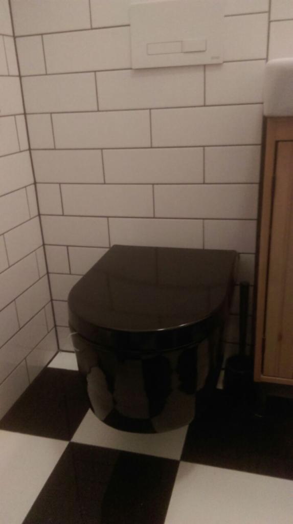 Czarna, podwieszana miska WC z deską wolnoopadającą, zamontowana na tle białych, prostokątnych płytek i czarno-białej podłogi w szachownicę, widoczny przycisk spłuczki.