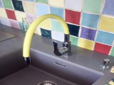 Bateria kuchenna z wylewką w jaskrawym żółtym oplocie na tle ściany z kolorowych płytek.