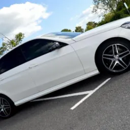 Biały samochód osobowy zaparkowany na parkingu z widocznym fragmentem nieba i drzew w tle.