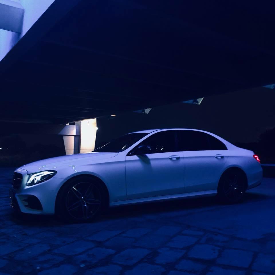 Biały samochód osobowy marki Mercedes-Benz klasy E zaparkowany pod betonowym wiaduktem w nocy, z widocznym oświetleniem ledowym i ciemnymi felgami.