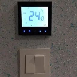 Czarny panel sterowania ogrzewaniem z wyświetlaną temperaturą 24 stopnie Celsjusza, zamontowany nad potrójnym, beżowym włącznikiem światła na ścianie o szarej, nakrapianej fakturze.