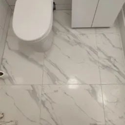 Biała toaleta, szafka i podłoga w łazience wyłożona białymi płytkami imitującymi marmur, widoczne pozostałości po remoncie przy wannie.