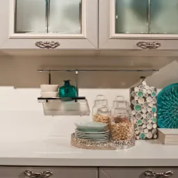 Dekoracyjna aranżacja na blacie kuchennym: szklane słoiki z orzechami, talerze na srebrnej tacy, turkusowa ozdobna misa i wazon z mozaiką, pod szafkami z matowymi szybami.