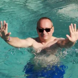 Mężczyzna w okularach przeciwsłonecznych, zanurzony w basenie, z wyciągniętymi rękami. Woda turkusowa, widoczne załamania światła.