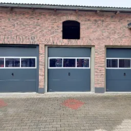 Trzy szare bramy garażowe segmentowe z oknami, w budynku z cegły klinkierowej z dekoracyjnym łukiem nad środkową bramą.