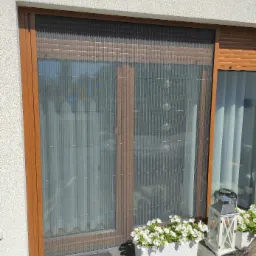 Zasłonięte moskitierą plisowaną brązowe okno w białym domu, obok okna roleta zewnętrzna, na zewnątrz donica z białymi kwiatami i dekoracyjny lampion.