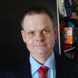 Portret mężczyzny w garniturze i czerwonym krawacie, z półuśmiechem, na tle szafy i półek z przedmiotami. Ujęcie od przodu, oświetlenie naturalne.