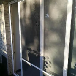 Biała rama okienna z siatką przeciw owadom, widoczne cienie liści na siatce, zewnętrzna ściana wyłożona płytkami imitującymi kamień.