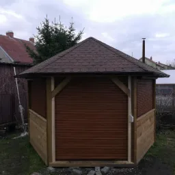 Altana ogrodowa z brązowymi rolowanymi osłonami okiennymi, drewnianą konstrukcją i dachem pokrytym gontem bitumicznym, na tle pochmurnego nieba.