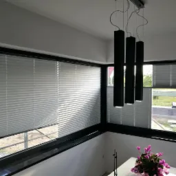 Wnętrze z oknami zabezpieczonymi plisowanymi osłonami przeciwsłonecznymi, widoczne czarne, wiszące lampy i kwiaty w wazonie.