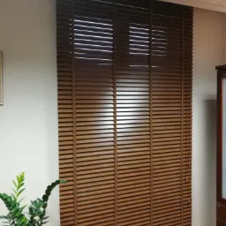 Brązowe drewniane żaluzje poziome zamontowane w oknie, częściowo podniesione, z widocznym mechanizmem regulacji sznurkowym, obok doniczka z zieloną rośliną.