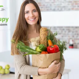 Uśmiechnięta kobieta trzyma brązową torbę z zakupami spożywczymi: pieczywo, warzywa, zieleń. W tle jasna kuchnia i logo firmy dostarczającej zakupy.