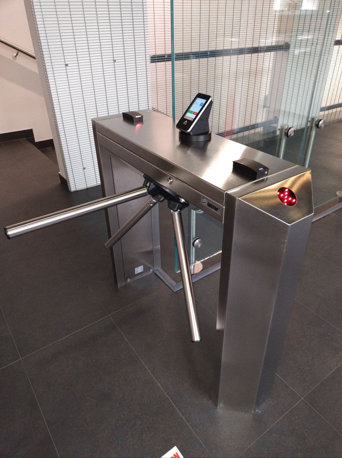 Metalowy kołowrót z czytnikiem biometrycznym i kontrolą dostępu, zainstalowany w nowoczesnym biurze z szklanymi ścianami i ciemną podłogą.