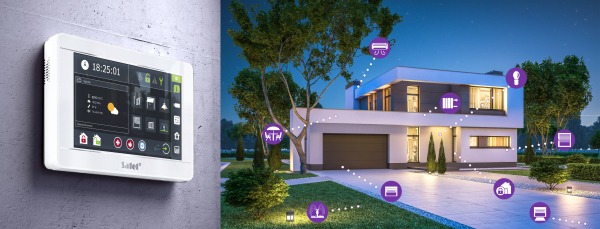 Panel sterowania systemem inteligentnego domu zamontowany na ścianie, z widokiem na nowoczesny dom z ogrodem oświetlonym nocą, ilustrujący zdalne zarządzanie funkcjami domu.