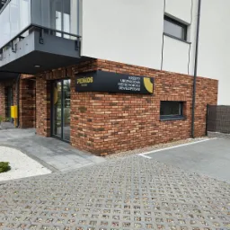 Budynek biurowy z cegły z szyldem informującym o kredytach, ubezpieczeniach nieruchomości i usługach deweloperskich, widok z zewnątrz na parking z ażurowej kostki.