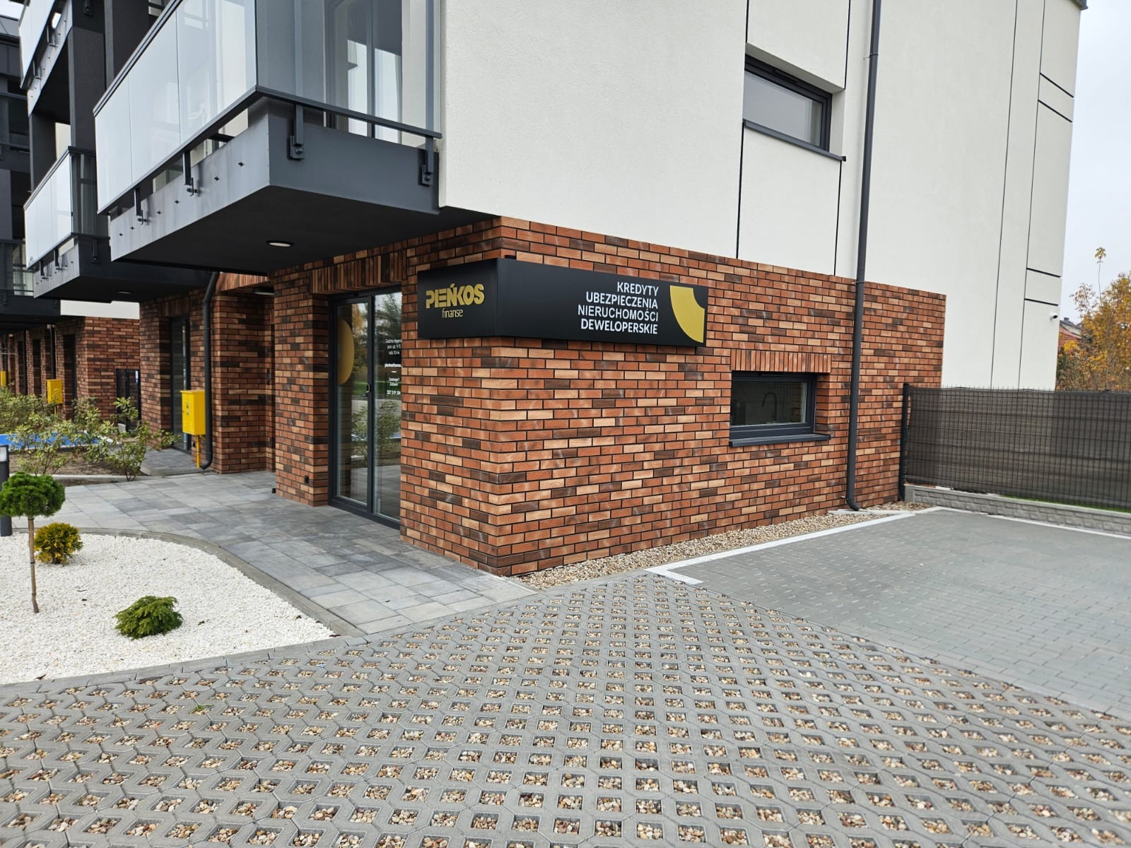 Budynek biurowy z cegły z szyldem informującym o kredytach, ubezpieczeniach nieruchomości i usługach deweloperskich, widok z zewnątrz na parking z ażurowej kostki.