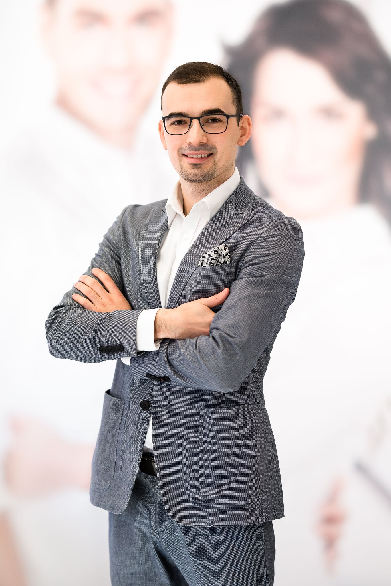 Uśmiechnięty mężczyzna w okularach i szarym garniturze z poszetką, stojący na tle rozmytego portretu dwóch osób.