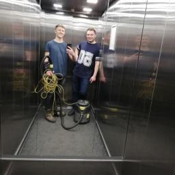 Nimboos - Dwaj mężczyźni w windzie, jeden trzyma zwinięty kabel, drugi robi selfie obok odkurzacza przemysłowego na dywanowej podłodze.