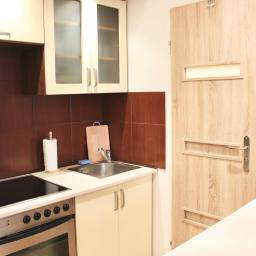 Nimboos - Niewielka, czysta kuchnia z beżowymi szafkami, brązowymi płytkami, płytą indukcyjną, piekarnikiem i czerwonym czajnikiem elektrycznym na białym blacie.