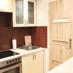 Niewielka, czysta kuchnia z beżowymi szafkami, brązowymi płytkami, płytą indukcyjną, piekarnikiem i czerwonym czajnikiem elektrycznym na białym blacie.