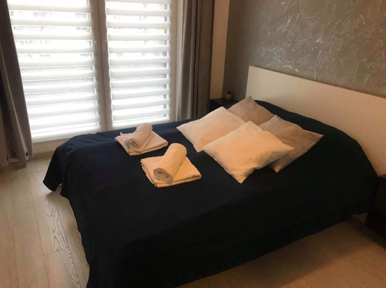 Uporządkowane łóżko z ciemnoniebieską narzutą, czterema poduszkami w odcieniach beżu i kremu oraz dwoma zestawami czystych, zwiniętych ręczników, na drewnianej podłodze, z widokiem na okno...