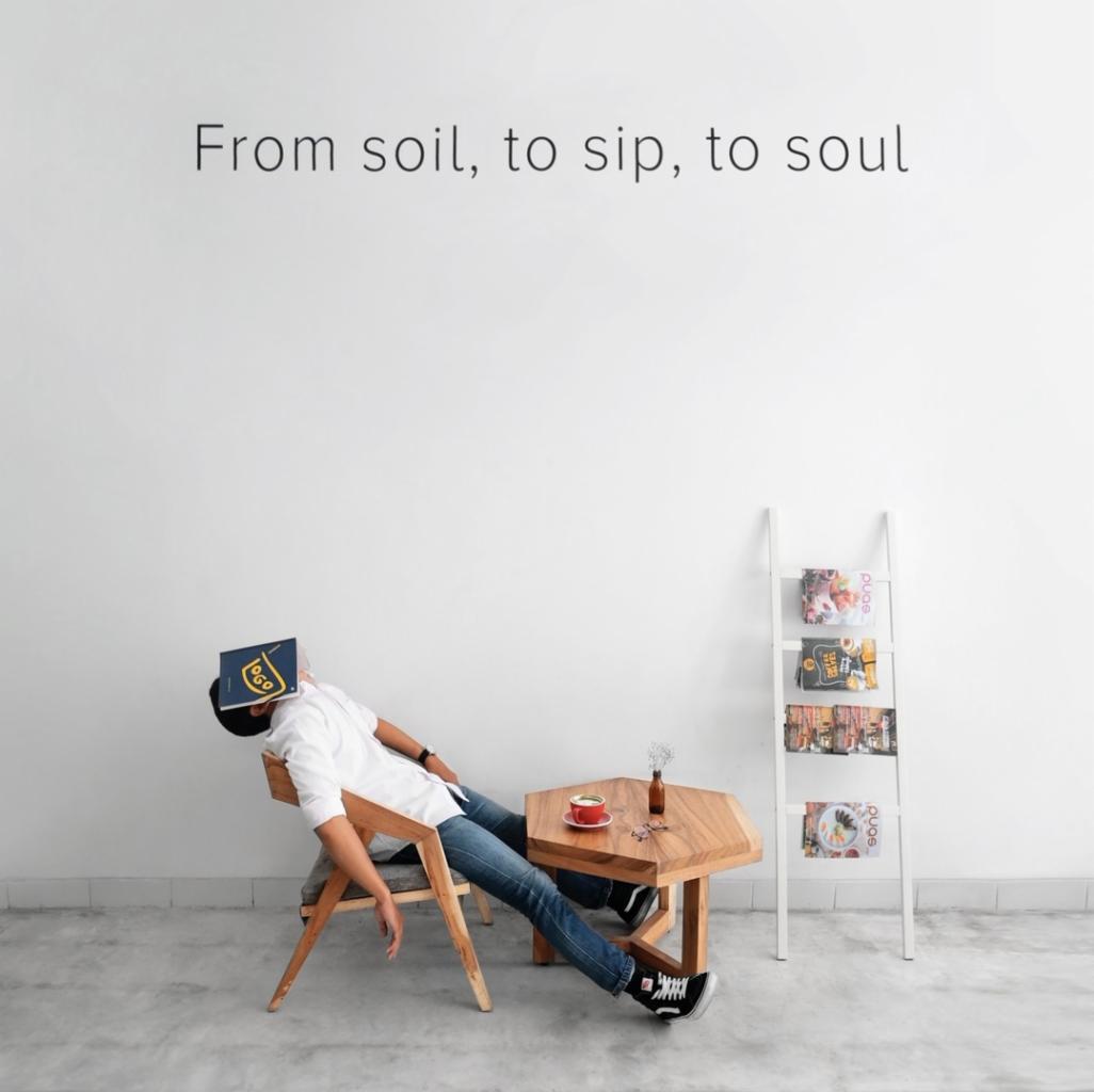Osoba w białej koszuli i dżinsach śpiąca na krześle z książką na twarzy, obok stolik z filiżanką i małą butelką, na tle białej ściany z napisem 'From soil, to sip, to soul' i białą drabinką...