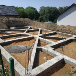 Zbrojone, betonowe fundamenty domu jednorodzinnego z wystającymi prętami zbrojeniowymi, widok z góry na wykop z ułożonym zbrojeniem. W tle widać dwa budynki w stanie surowym otwartym i pryzmę ziemi.
