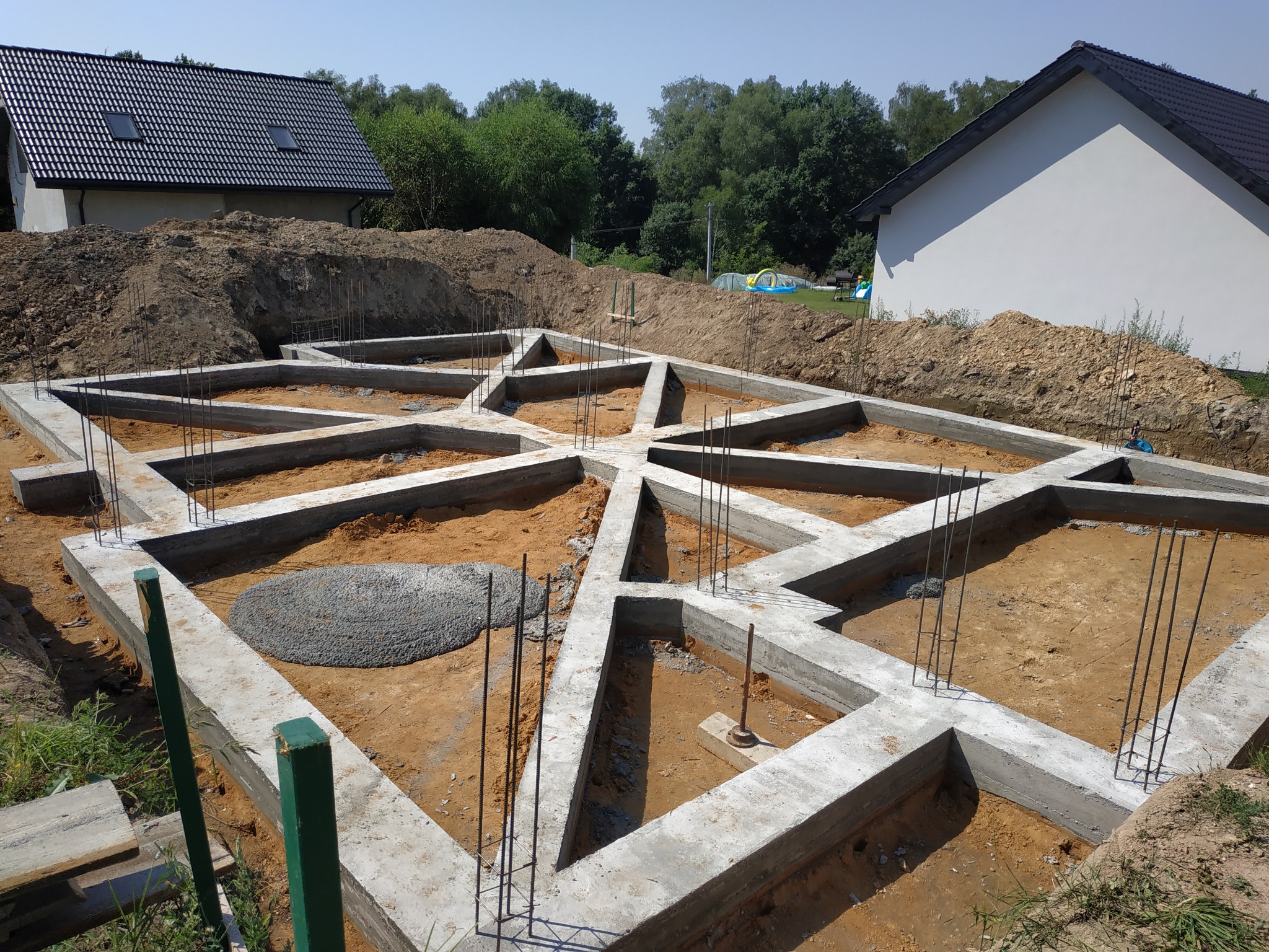 Zbrojone, betonowe fundamenty domu jednorodzinnego z wystającymi prętami zbrojeniowymi, widok z góry na wykop z ułożonym zbrojeniem. W tle widać dwa budynki w stanie surowym otwartym i pryzmę ziemi.