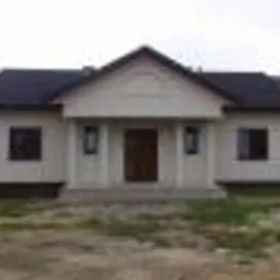 Gotowy, parterowy dom z jasną elewacją i ciemnym dachem, widoczny z przodu, z zielonym trawnikiem i fragmentem sąsiedniego budynku w tle. Prosta bryła, symetryczny układ.