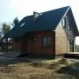 Gotowy, brązowy dom z ciemnym dachem, z werandą, na działce w trakcie zagospodarowywania, w słoneczny dzień.