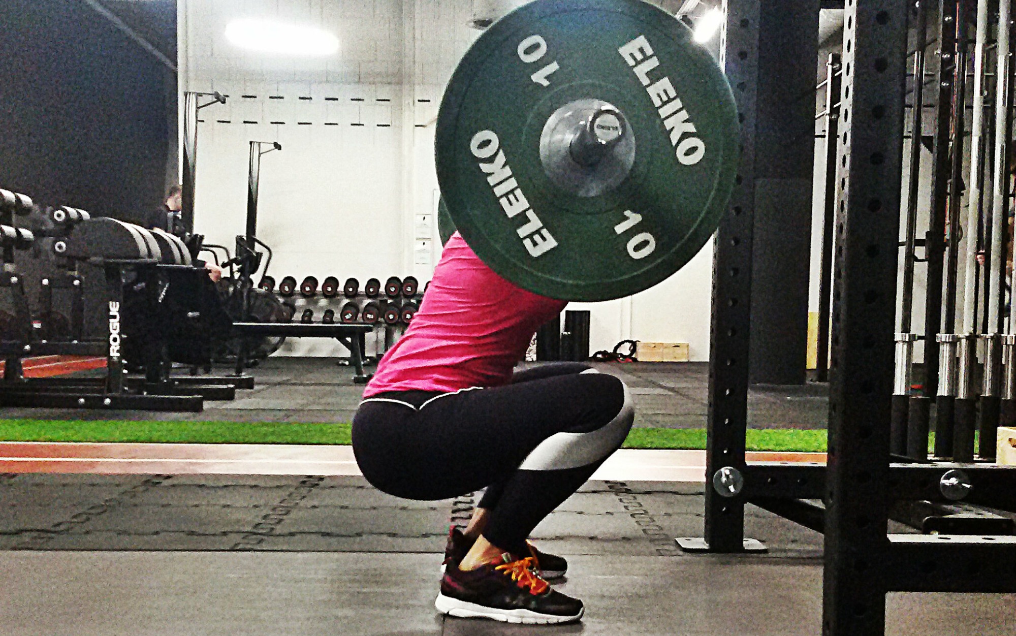 Kobieta w różowej koszulce wykonuje przysiad ze sztangą Eleiko 10kg w siłowni; widoczny sprzęt Rogue i regał z hantlami w tle.