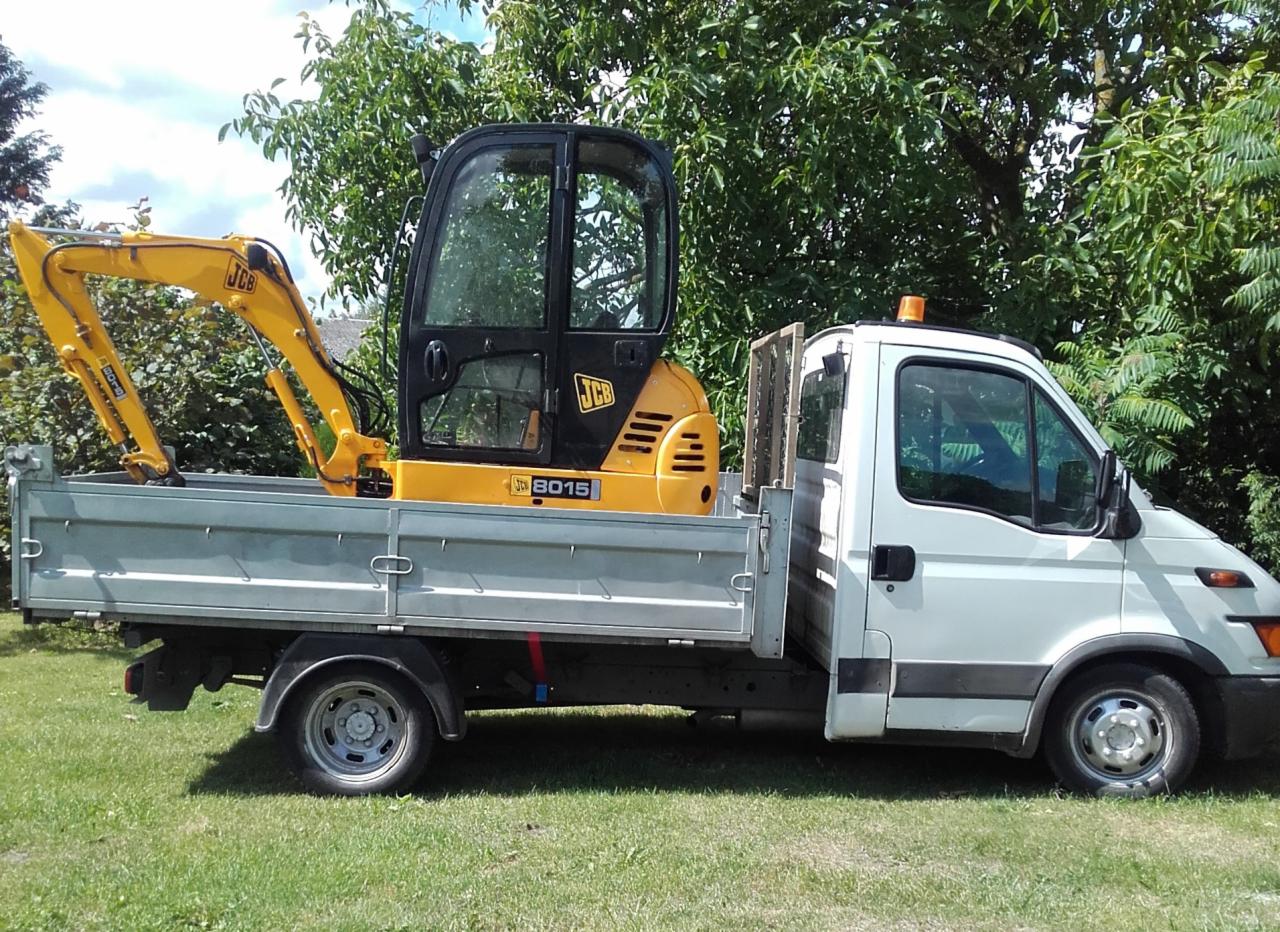 Żółta mini koparka JCB 8015 przewożona na platformie białego samochodu dostawczego na tle zieleni.