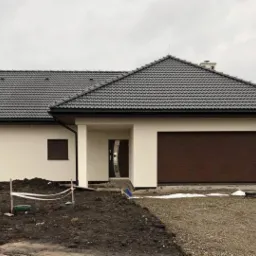 Nowoczesny dom jednorodzinny z brązową bramą garażową segmentową, brązowymi roletami zewnętrznymi i ciemnoszarą dachówką, widok z zewnątrz w pochmurny dzień.