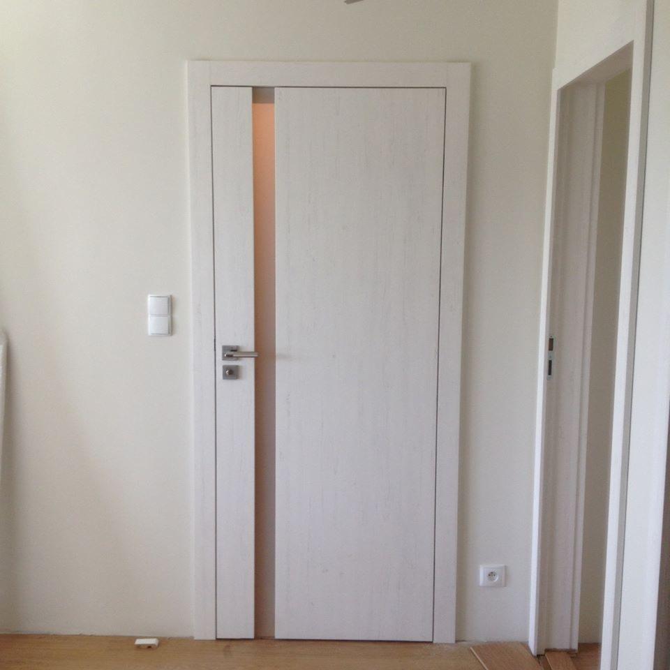 Białe drzwi wewnętrzne z wąskim pionowym przeszkleniem, osadzone w białej framudze, widoczne na tle jasnej ściany i podłogi, obok widoczny fragment drugich drzwi.