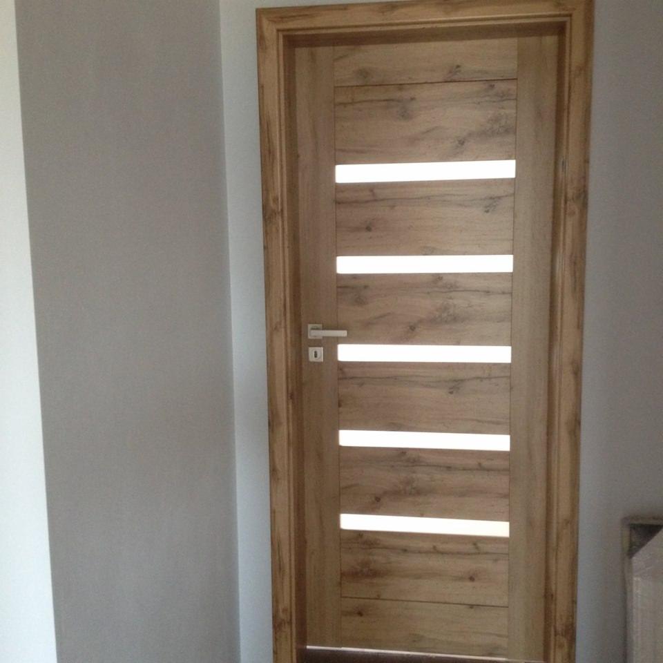 Drewniane drzwi wewnętrzne z poziomymi, podświetlanymi paskami, zamontowane w szarej ścianie.