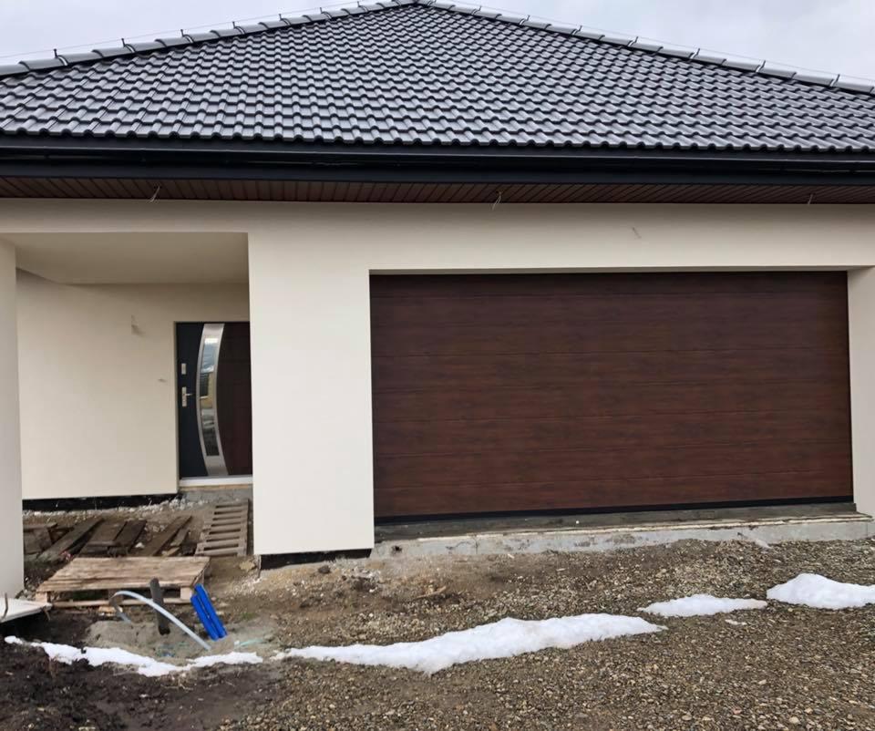 Nowoczesny dom z brązową bramą garażową segmentową o fakturze drewna i drzwiami wejściowymi z elementami stali nierdzewnej, widoczne ślady prac wykończeniowych na zewnątrz.