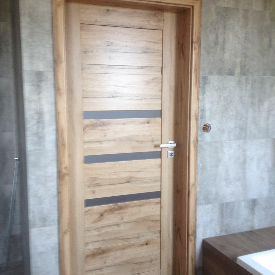 Jasne, drewnopodobne drzwi wewnętrzne z poziomymi, szarymi wstawkami, zamontowane w łazience z szarymi płytkami.