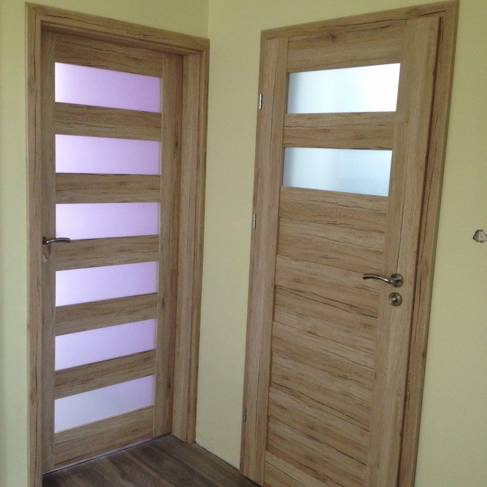 Dwa drewniane drzwi wewnętrzne z przeszkleniami: jedne z czterema poziomymi szybami w odcieniach fioletu, drugie z trzema szybami mlecznymi, zamontowane w jasnym pomieszczeniu z drewnianą podłogą.