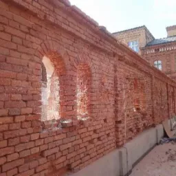 Ceglana ściana budynku z widocznymi śladami erozji i ubytkami w spoinach, z oknami w kształcie łuków, w tle budynek w podobnym stylu architektonicznym.