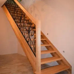 Jasne drewniane schody z ozdobną, kutą balustradą w nowoczesnym wnętrzu z beżowymi płytkami podłogowymi i ścianami w odcieniach beżu i bieli.