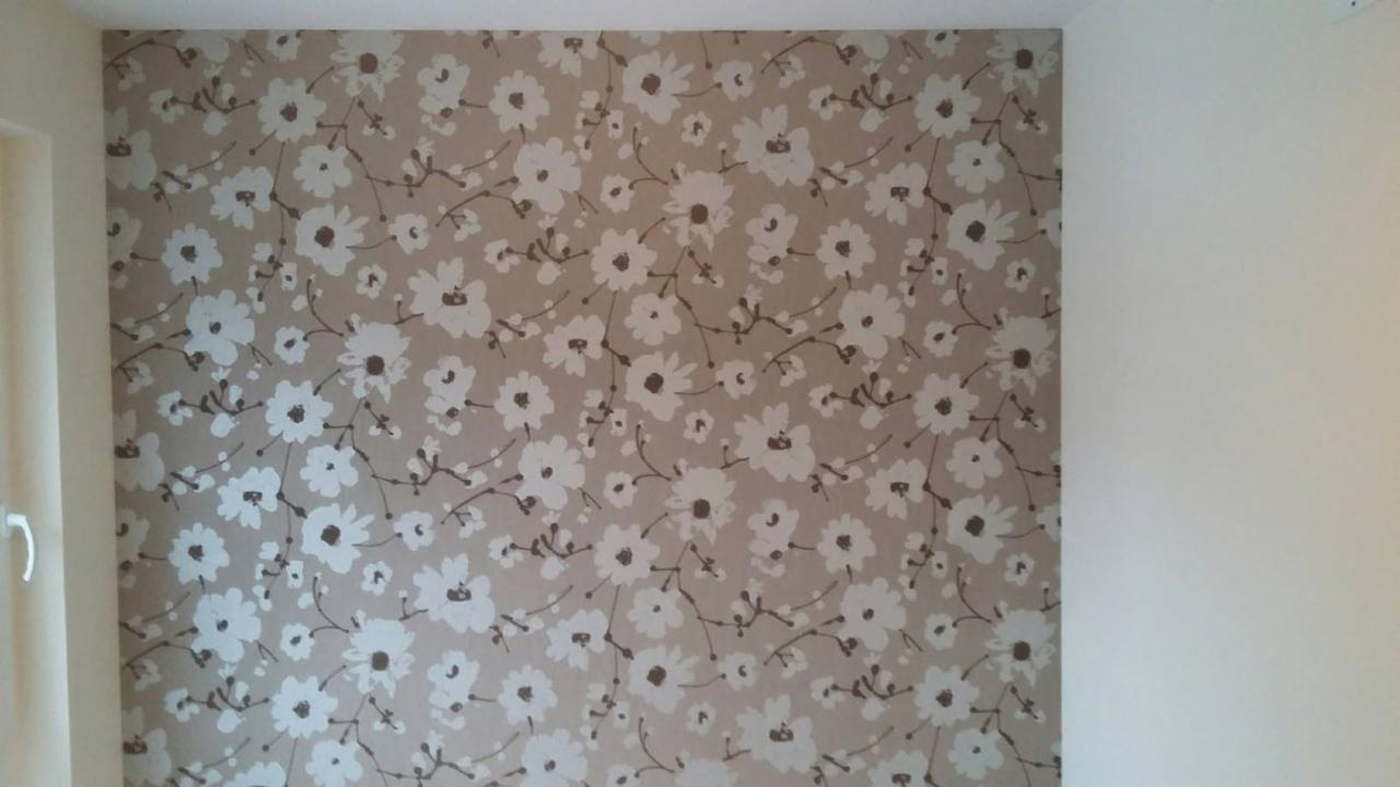 Ściana pokryta beżową tapetą w białe kwiaty z brązowymi środkami, widok z bliska na wzór, z fragmentem białej ściany po prawej stronie i ościeżnicą okna po lewej.