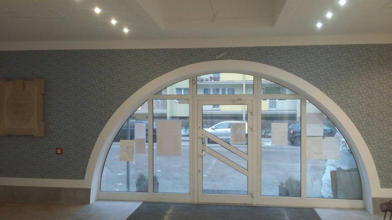 Ściana pokryta tapetą w odcieniach szarości z delikatnym wzorem, przylegająca do łukowego okna/drzwi wejściowych z białym obramowaniem, widoczne detale wykończeniowe i oświetlenie sufitowe.