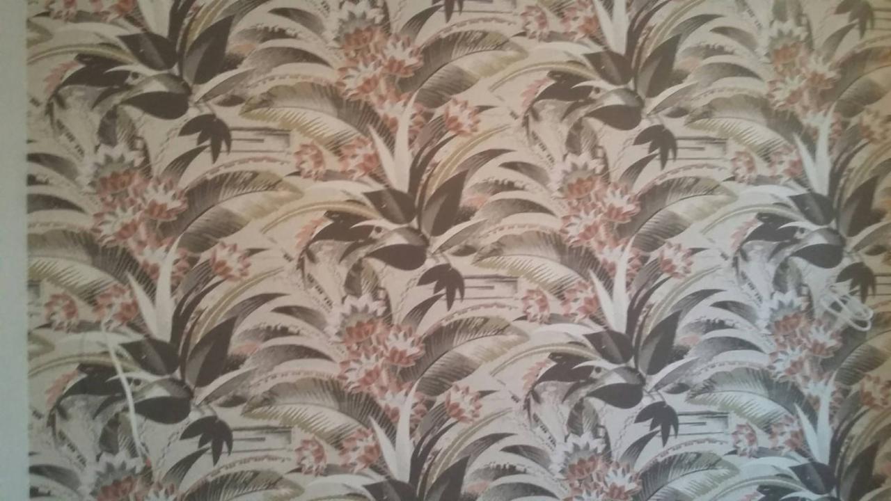 Fragment ściany pokrytej tapetą z wzorem tropikalnych liści palmowych i kwiatów w odcieniach brązu, beżu i delikatnego różu.