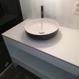 Nowoczesna, okrągła umywalka nablatowa w kolorze czarno-białym, zamontowana na białej szafce z półkami, z widocznym czarnym kranem i gniazdkiem elektrycznym na ścianie w tle.