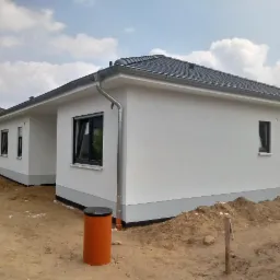 Nowo wybudowany, parterowy dom z białymi ścianami i szarą dachówką, widoczny z zewnątrz, z pomarańczowymi elementami kanalizacji w ziemi.