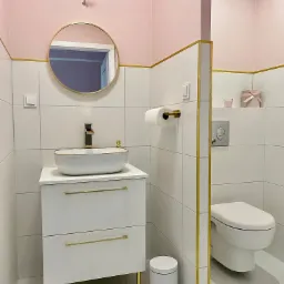 Nowoczesna łazienka w bieli i różu z złotymi akcentami: umywalka nablatowa, okrągłe lustro, biała toaleta i dekoracyjne pudełko na półce.